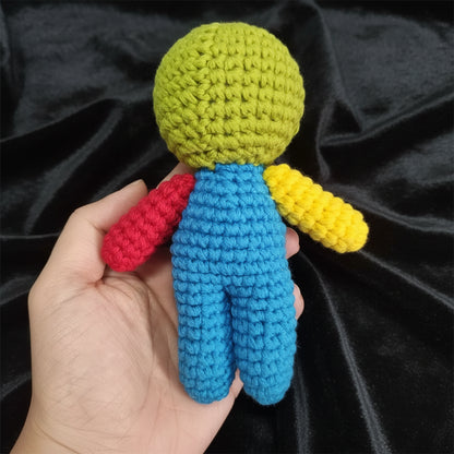 A small crocheted doll held in a hand, featuring a lime green head, blue body, one red arm, and one yellow arm, against a plain light gray background.