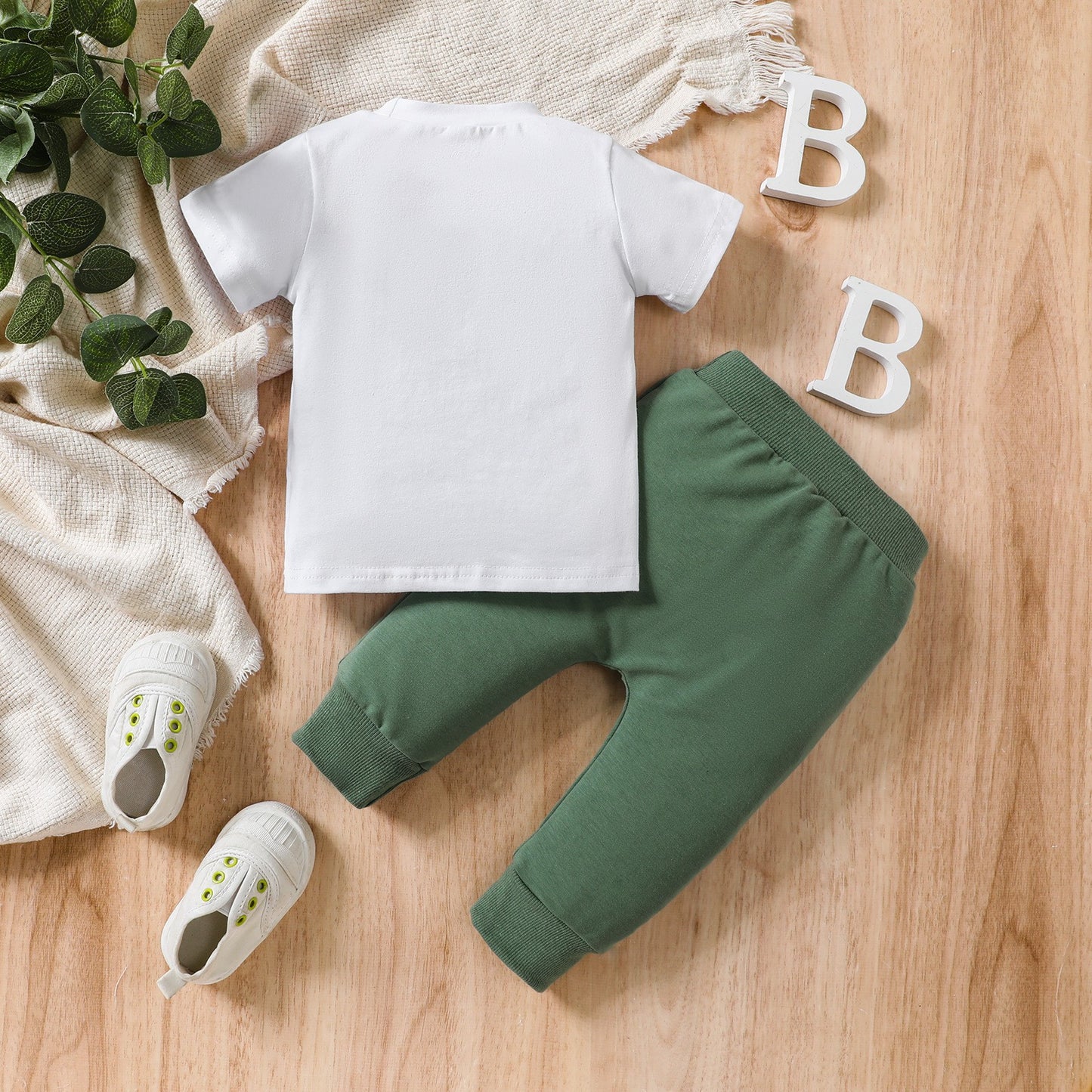 White t-shirt, green pants, and baby shoes on a wooden surface with decorative elements.