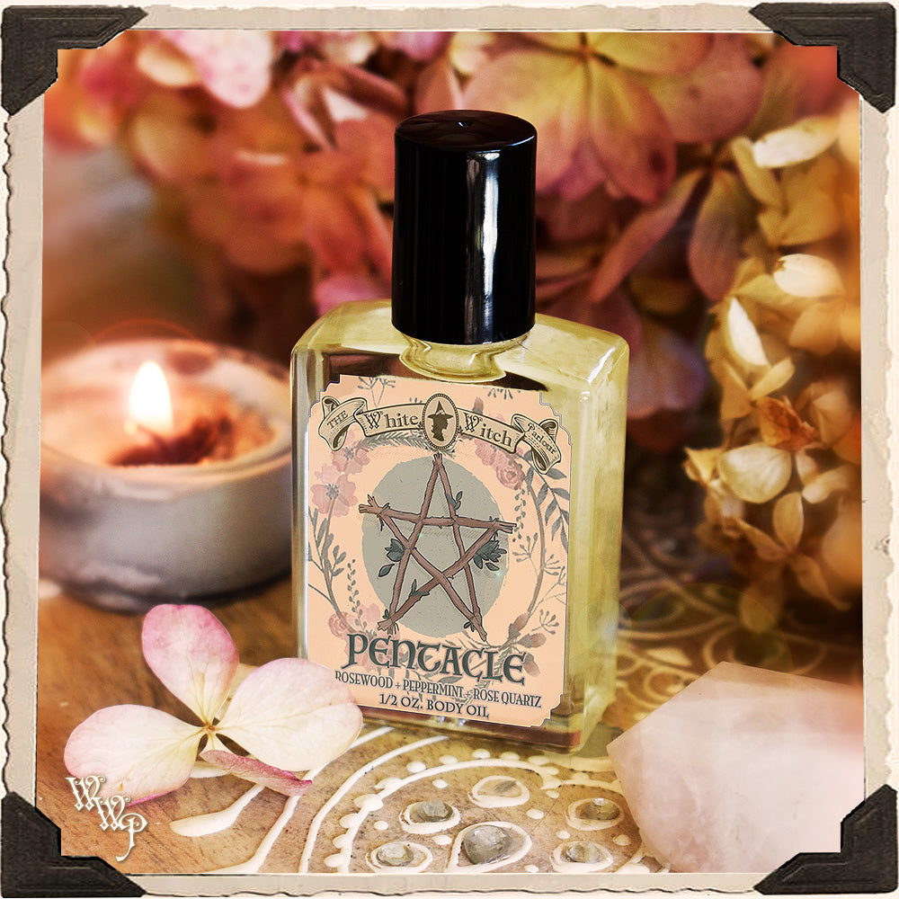 Body oil bottle with pentacle design on label, surrounded by flowers and a candle.