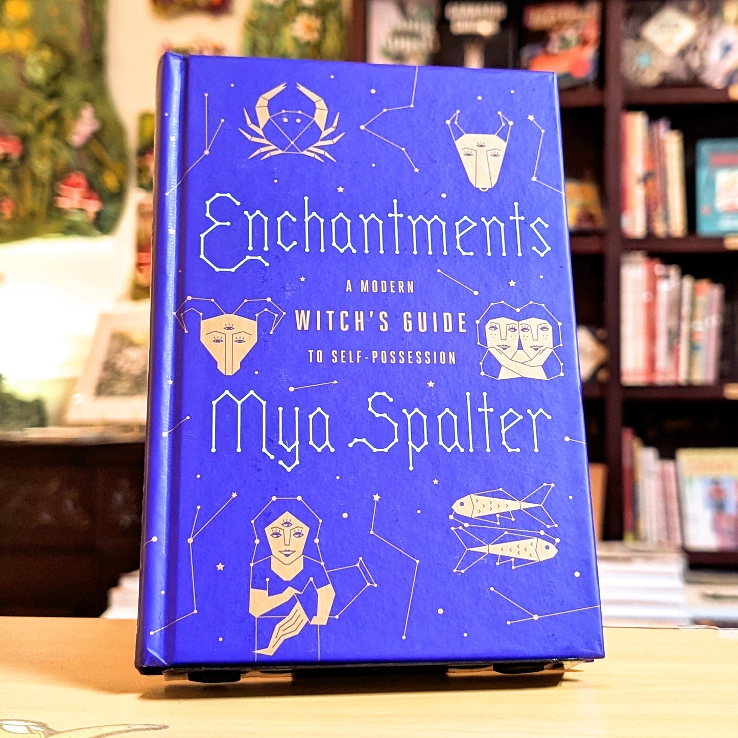 Blue book titled 'Enchantments' by Mya Spalter on a wooden surface with a blurred bookshelf background.