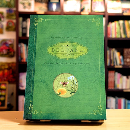Green book titled 'Beltane' with decorative elements on a wooden surface and bookshelf background.