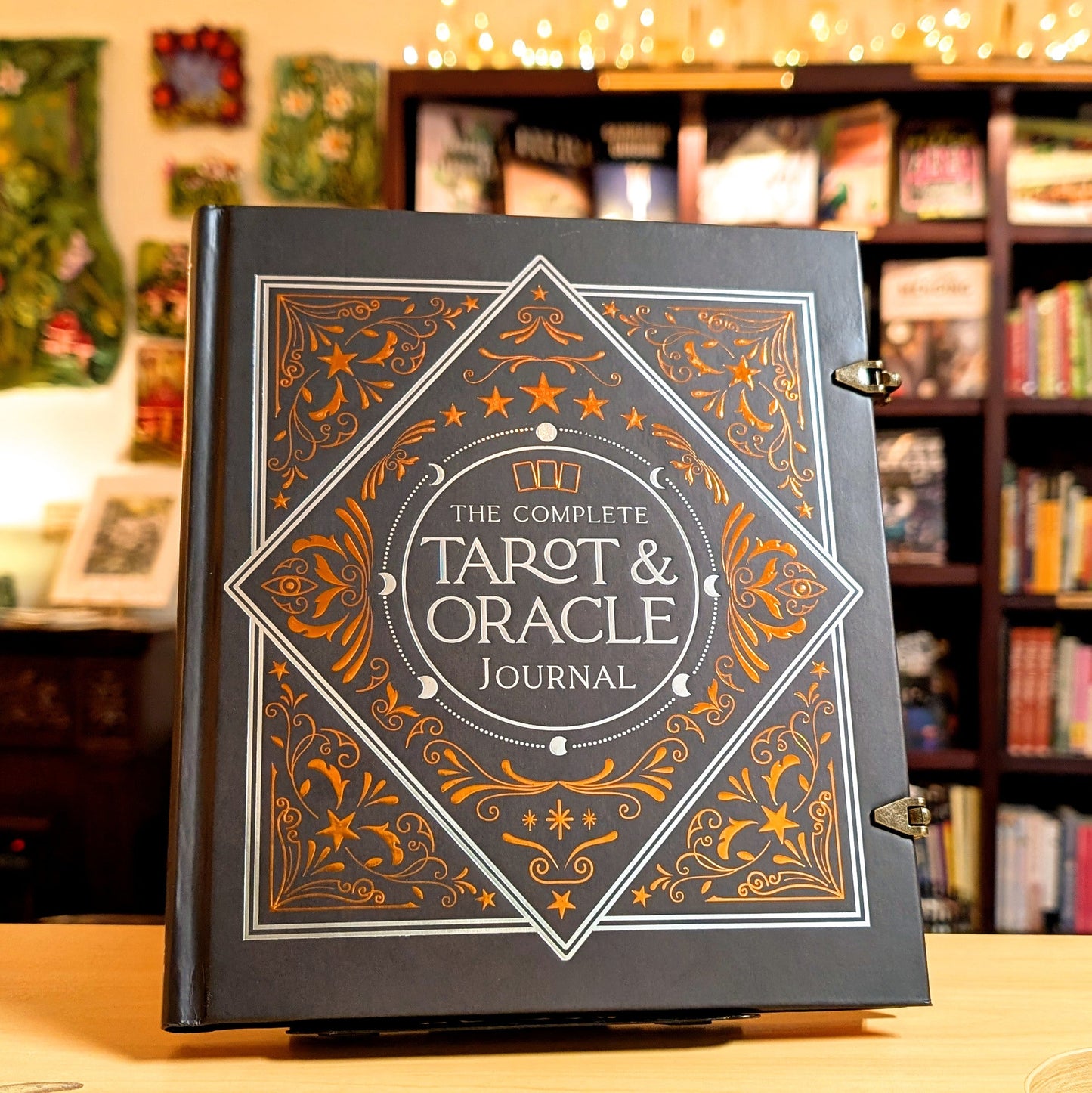 The Complete Tarot & Oracle Journal standing upright on a wooden table, with a blurred library or study background featuring bookshelves and warm string lights.