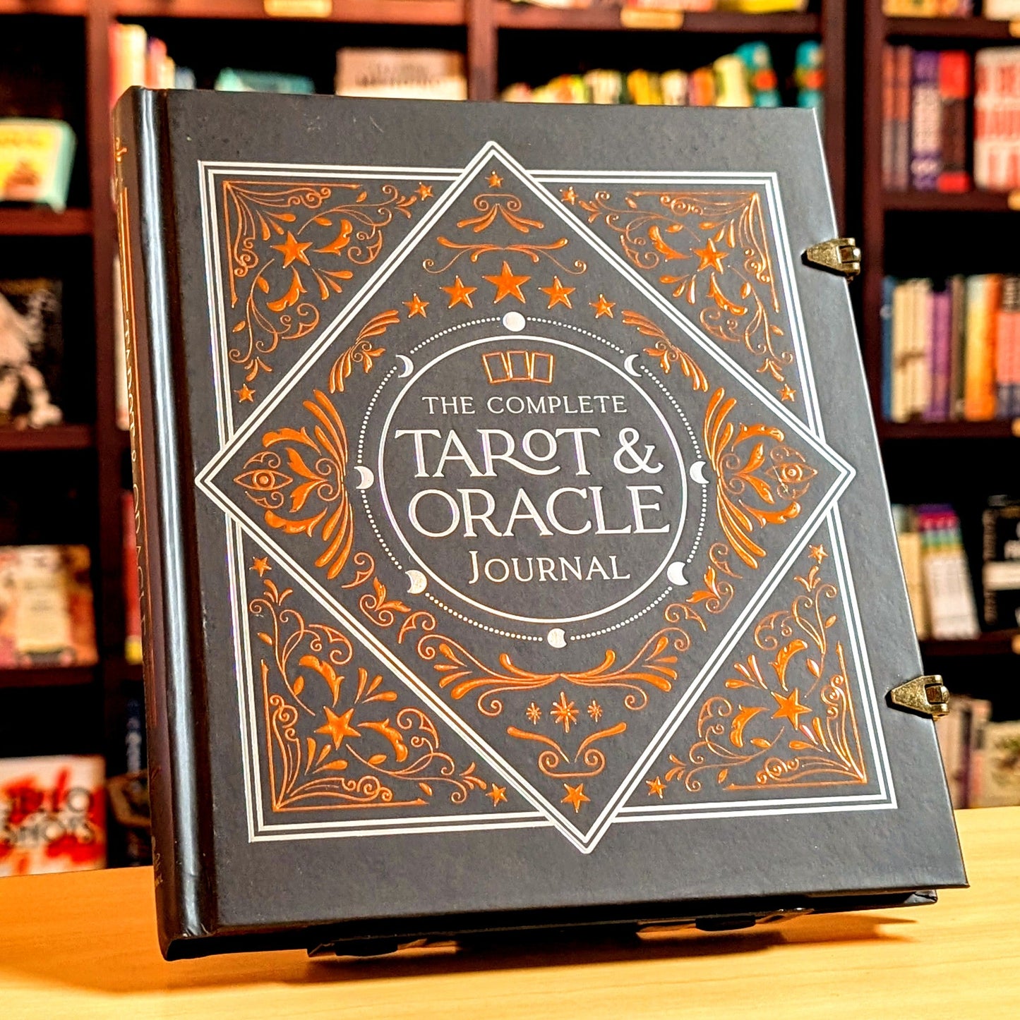 The Complete Tarot & Oracle Journal standing upright on a wooden table with a blurred bookshelf background. The journal has a dark gray cover with ornate gold and white gothic-style illustrations and lettering.