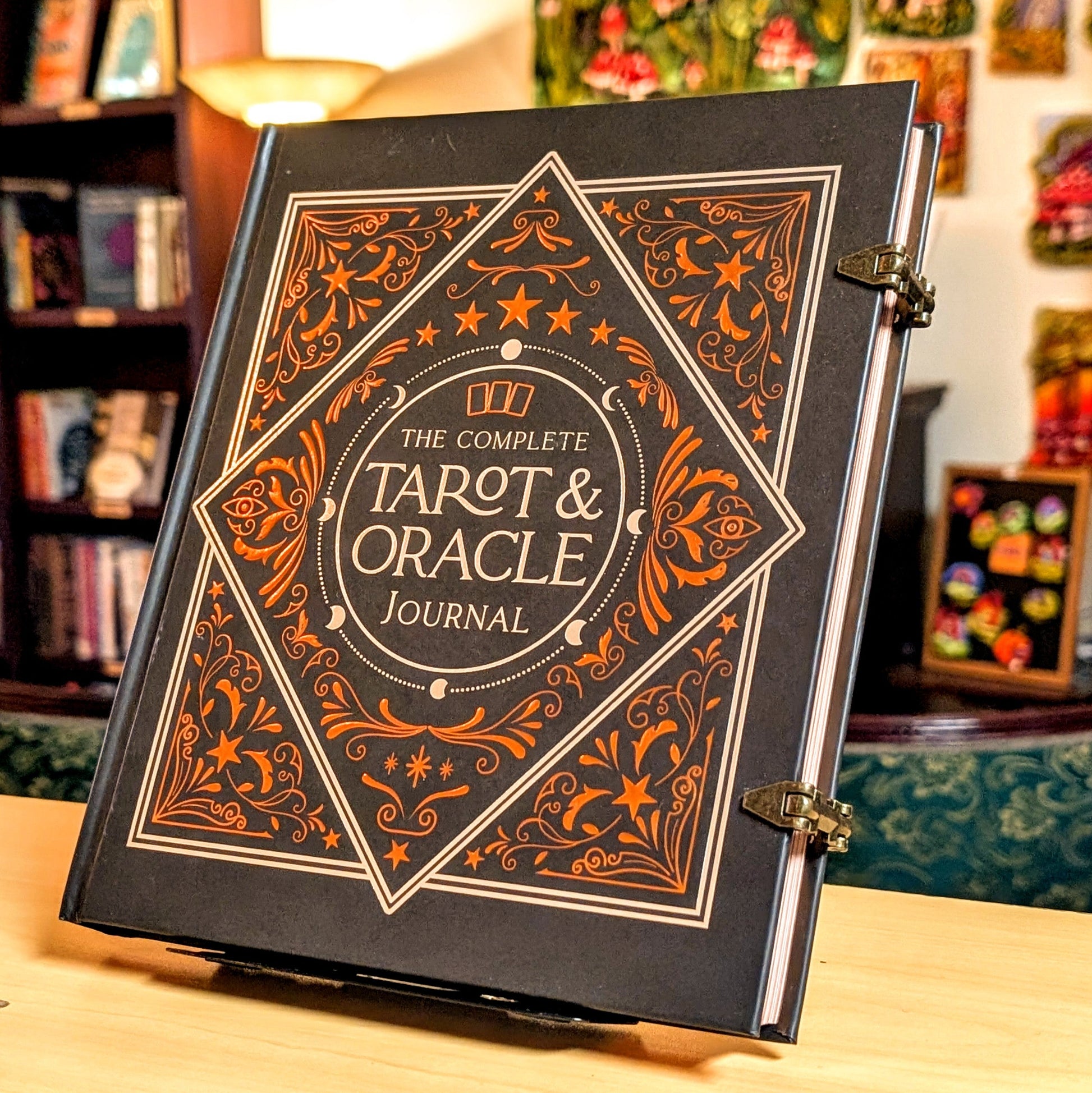 The Complete Tarot & Oracle Journal standing upright on a wooden surface in a cozy, decorated room. The book cover is black with intricate orange and gold filigree patterns and gold lettering.