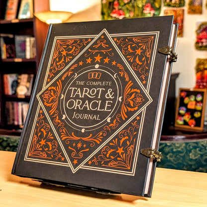 The Complete Tarot & Oracle Journal standing upright on a wooden surface in a cozy, decorated room. The book cover is black with intricate orange and gold filigree patterns and gold lettering.