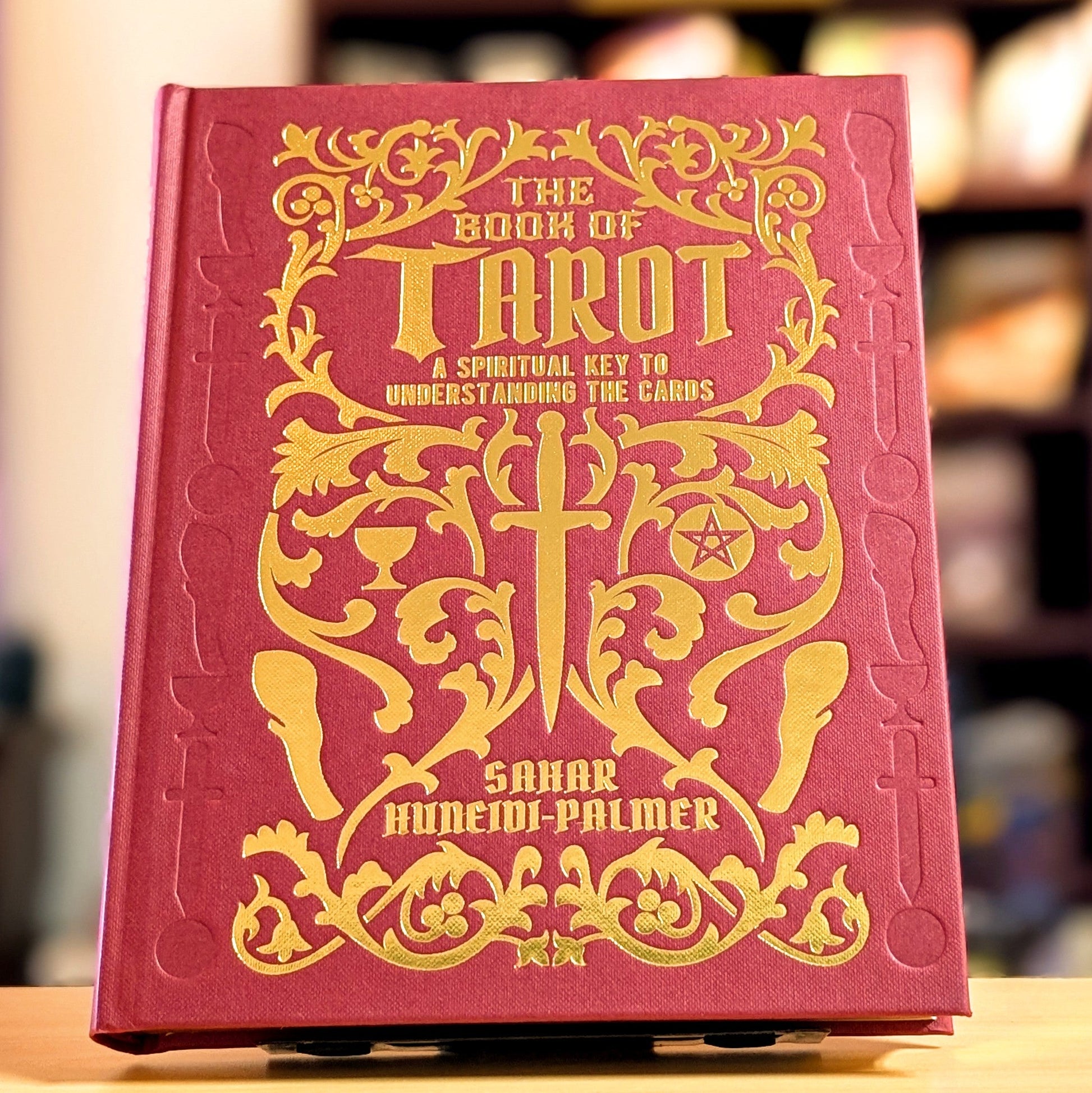 Red book titled 'The Book of Tarot' with gold embossed text on a blurred bookshelf background.