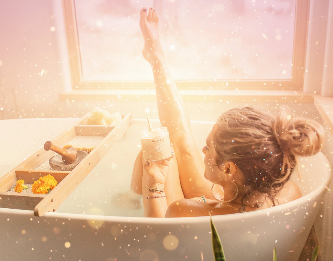 Woman relaxing in a bathtub with a drink, surrounded by bath bombs and oils.