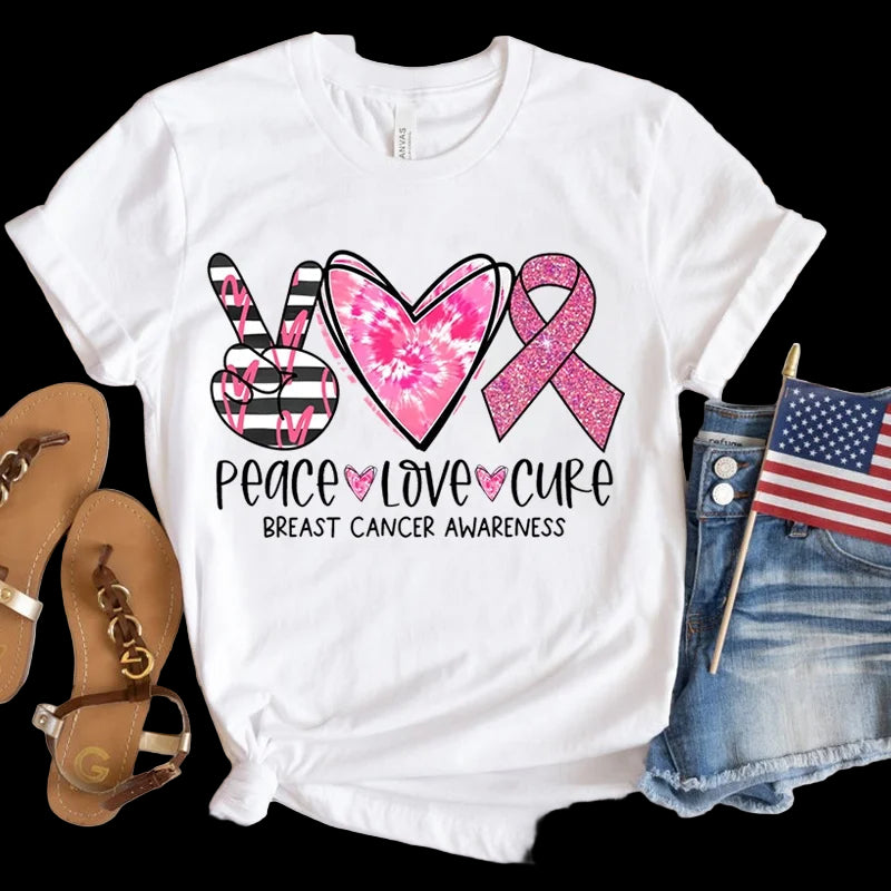 White t-shirt with pink breast cancer awareness design on a wooden surface with sandals and denim shorts.