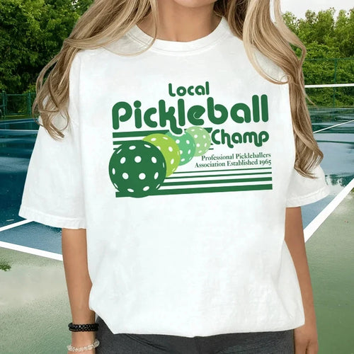 Person wearing a white t-shirt with pickleball-themed design on a tennis court.