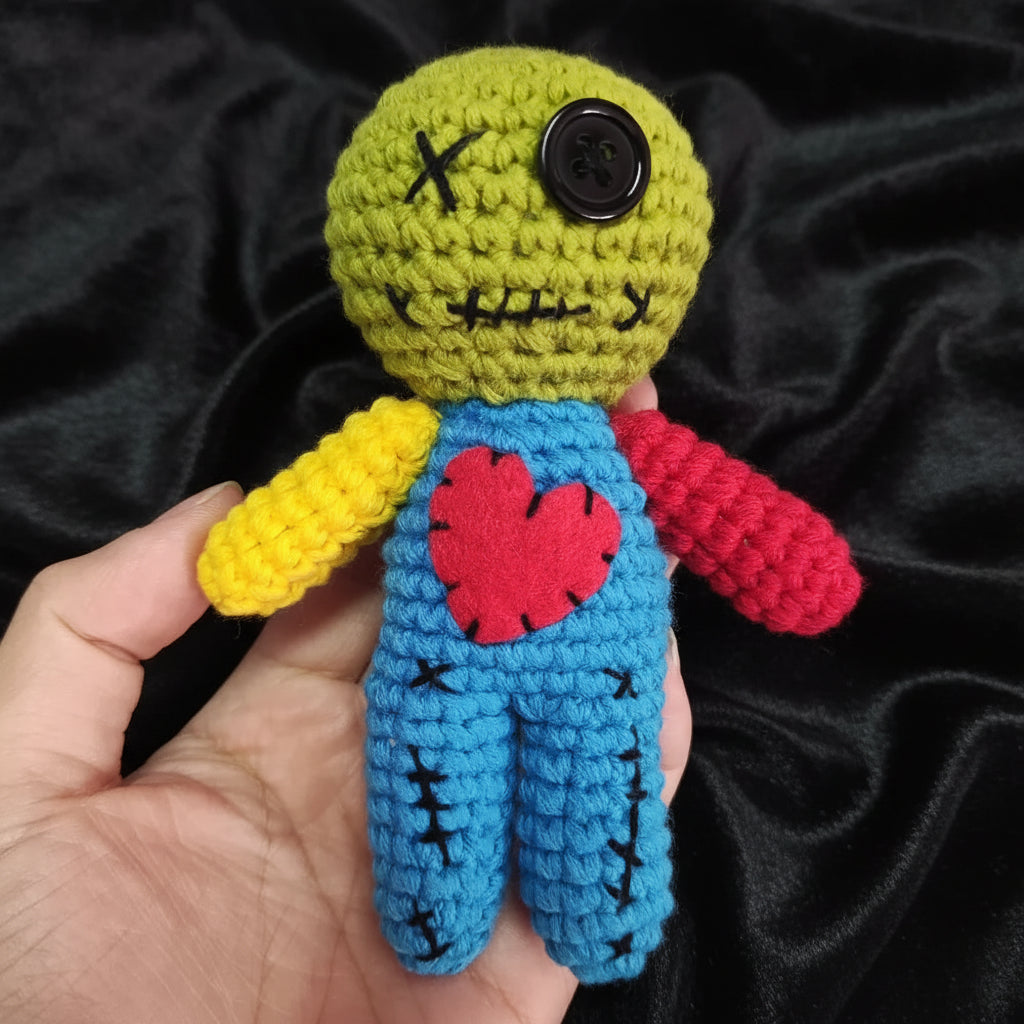Handmade crochet toy with green head, blue body, yellow arms, pink shirt, and red heart on a light gray background.