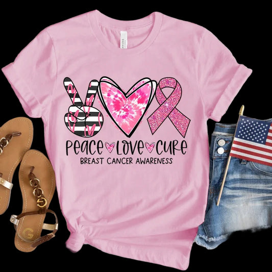 Pink t-shirt with peace, love, cure design for breast cancer awareness on a wooden surface with sandals and an American flag.