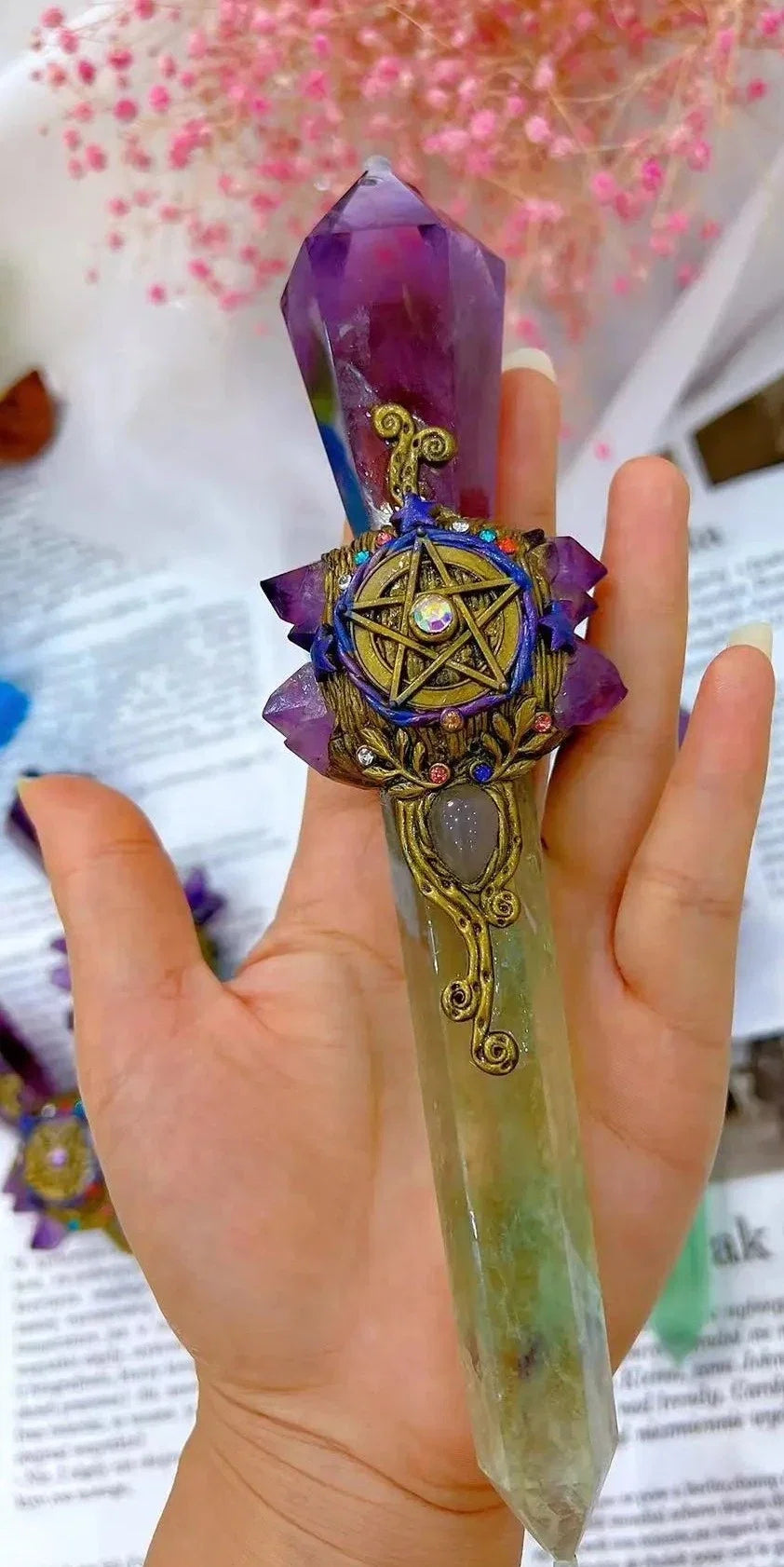 A hand holding a decorative crystal wand with a purple amethyst top, a green and white chakra stone midsection, and an ornate gold-toned pentacle centerpiece with purple petals.