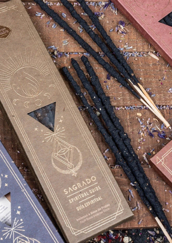 Sacrado spiritual incense sticks with black-coated wicks on wooden sticks, displayed next to their brown kraft paper packaging box on a rustic wooden surface with dried lavender petals.