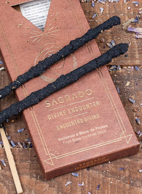 Two black ceremonial drug sticks from the brand Sacrado lie on top of their brown cardboard packaging, which is placed on a rustic wooden surface scattered with dried lavender.