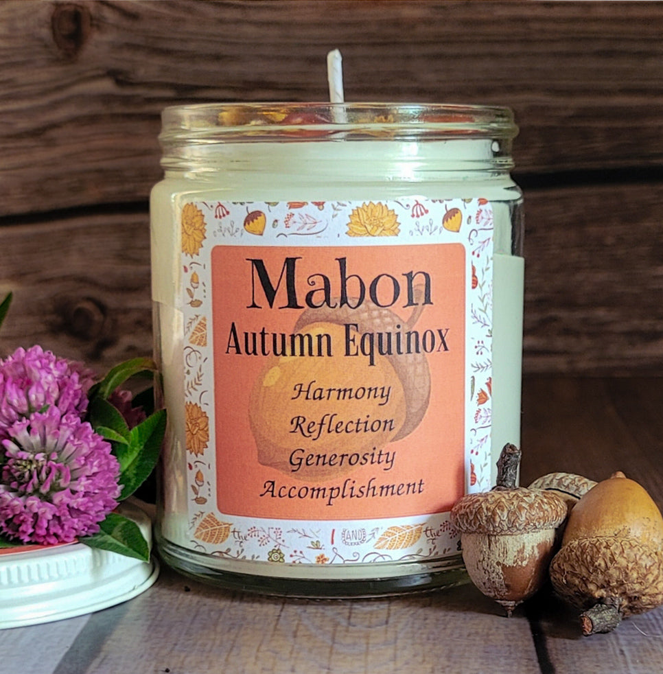 A glass jar candle labeled 'Cabon Autumn Equinox' with a white wax fill and a peach-colored label featuring autumn leaf illustrations. The candle is styled on a rustic wooden surface with acorns and a purple flower in the foreground.