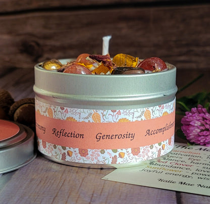 A round metal candle tin filled with amber-colored wax and decorative stones, set on a wooden surface with a floral label reading 'Reflection Generosity Accomplishment'.