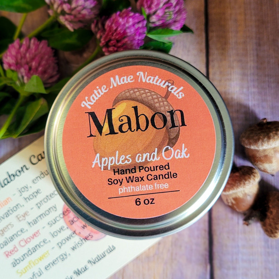A round metal tin of Mabon hand-poured soy wax candle by Katie Mae Naturals, scented with apples and oak, shown on a rustic wooden surface with purple flowers and acorns around it.