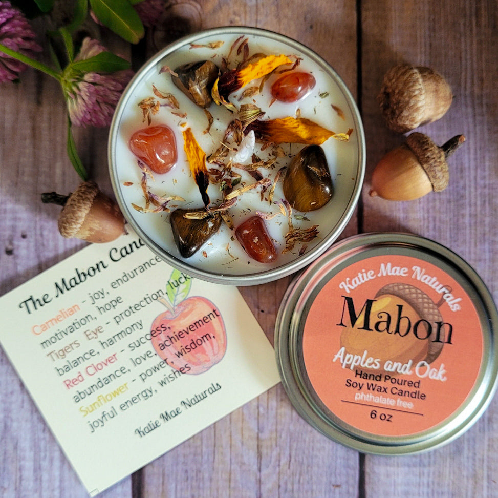An open round metal tin of Mabon soy wax candle with visible botanical wax pieces and dried flower petals, shown beside the matching lid on a rustic wood surface. A printed candle card for 'The Mabon Candle Co.