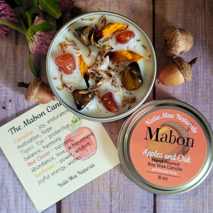 An open round metal tin of Mabon soy wax candle with visible botanical wax pieces and dried flower petals, shown beside the matching lid on a rustic wood surface. A printed candle card for 'The Mabon Candle Co.