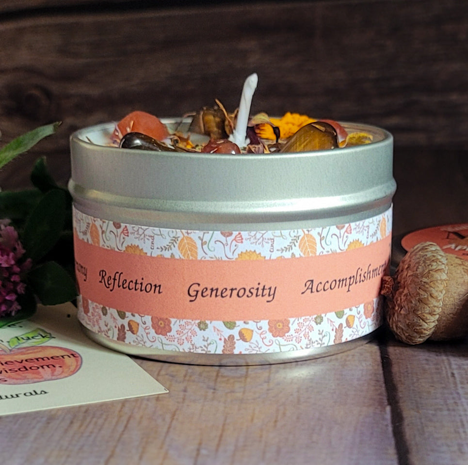 A round metal tin candle with a floral label reading 'Reflection, Generosity, Accomplishment' sits on a wooden surface with dried flowers and a wick visible inside. The scene is styled with autumn-themed props like a pinecone and leaves.