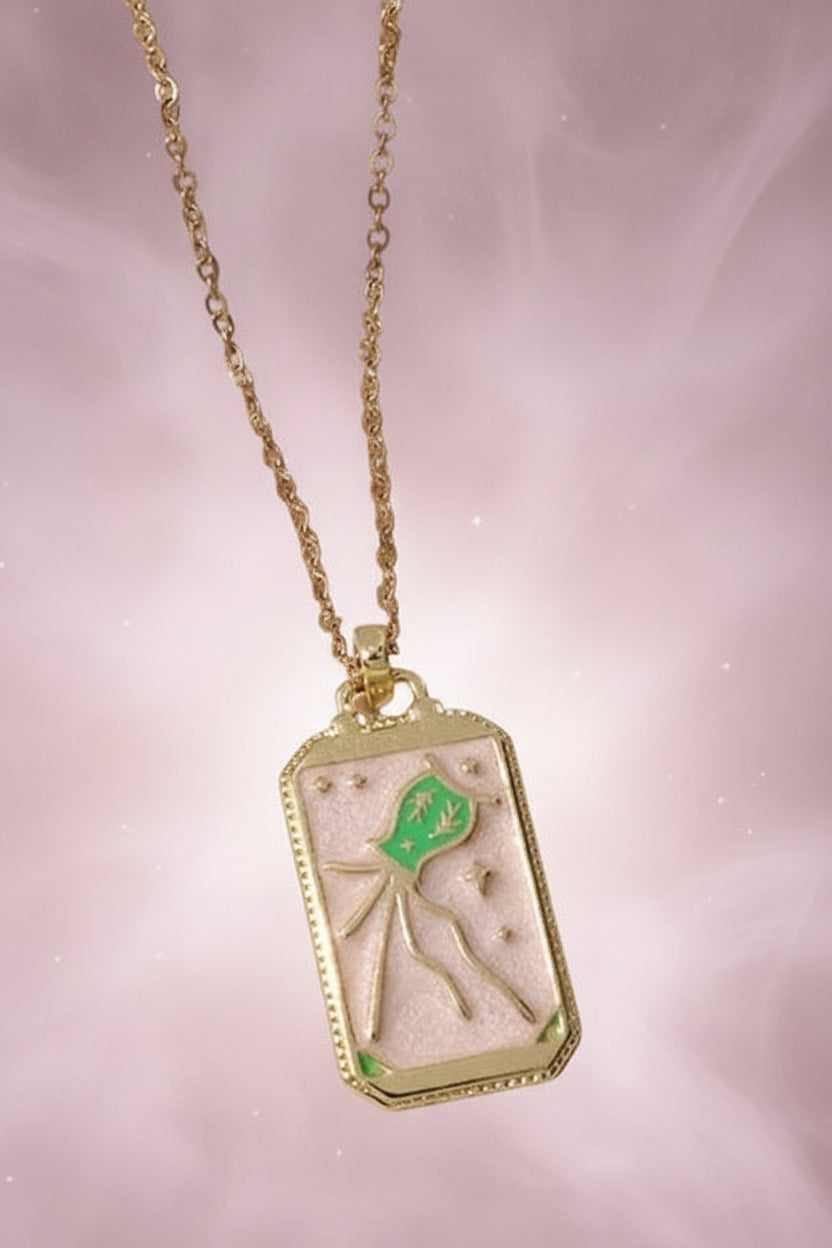 Gold necklace with a rectangular pendant featuring a green leaf on a textured beige surface