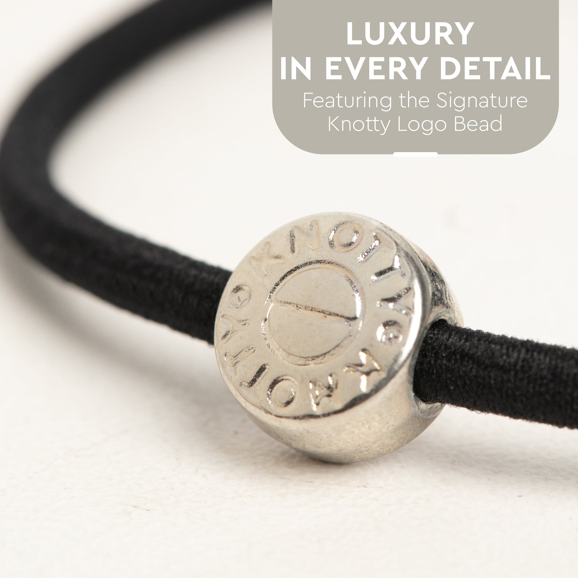 Silver bead with branding on a black cord against a white background, featuring text about luxury and the signature knotty logo bead.
