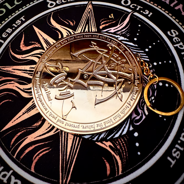 A circular metallic pendant or medallion with engraved script and a pentagram design, attached to a gold key ring, displayed on a black occult-style printed background with celestial symbols.