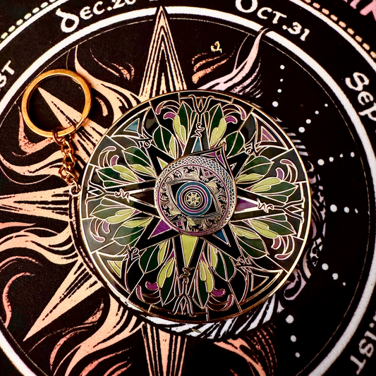 A circular enamel pin with a compass rose design and a gold keyring, displayed on a black circular background with ornate gold illustrations.