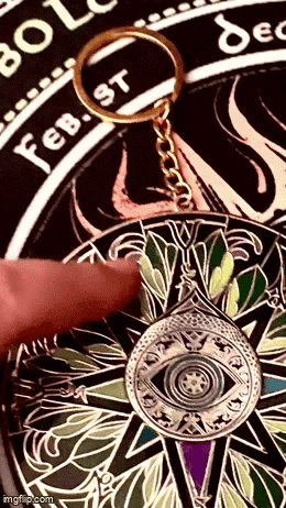 A close-up of a decorative keychain featuring a silver-toned eye motif and gold key ring, resting on a black circular card with white text and green, purple, and gold illustrated details. A finger points toward the central eye design.