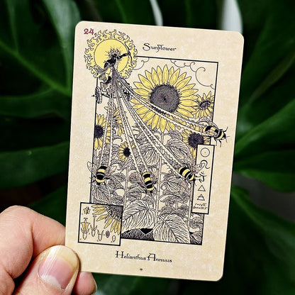 A hand holding a tarot-style card featuring a sunflower illustration, with green leaves blurred in the background.
