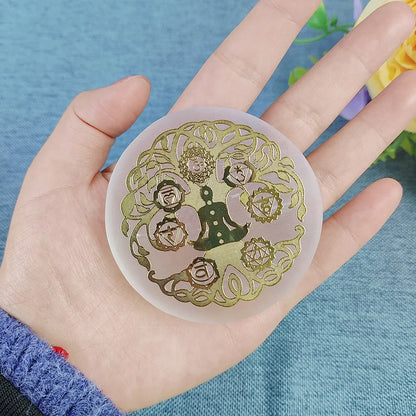 A hand holding a round translucent silicone mold or coaster featuring a gold foil design of a figure meditating within a floral and knot pattern, set against a blue textured background.