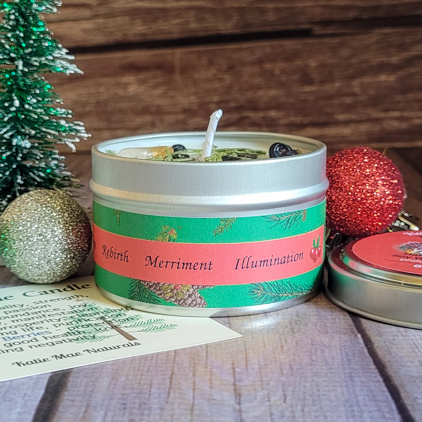 Candle in a tin with decorative label surrounded by Christmas ornaments on a wooden surface