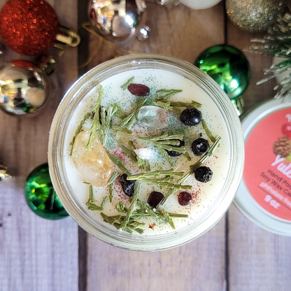 Jar of candle with herbs and berries on a wooden surface with Christmas ornaments.