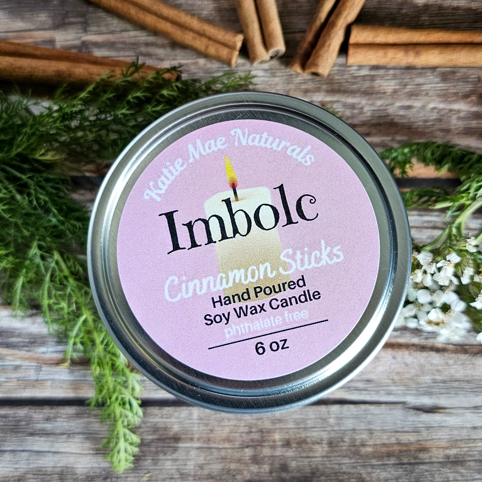 Candle jar labeled 'Imbolc Cinnamon Sticks' with cinnamon sticks and greenery on a wooden surface