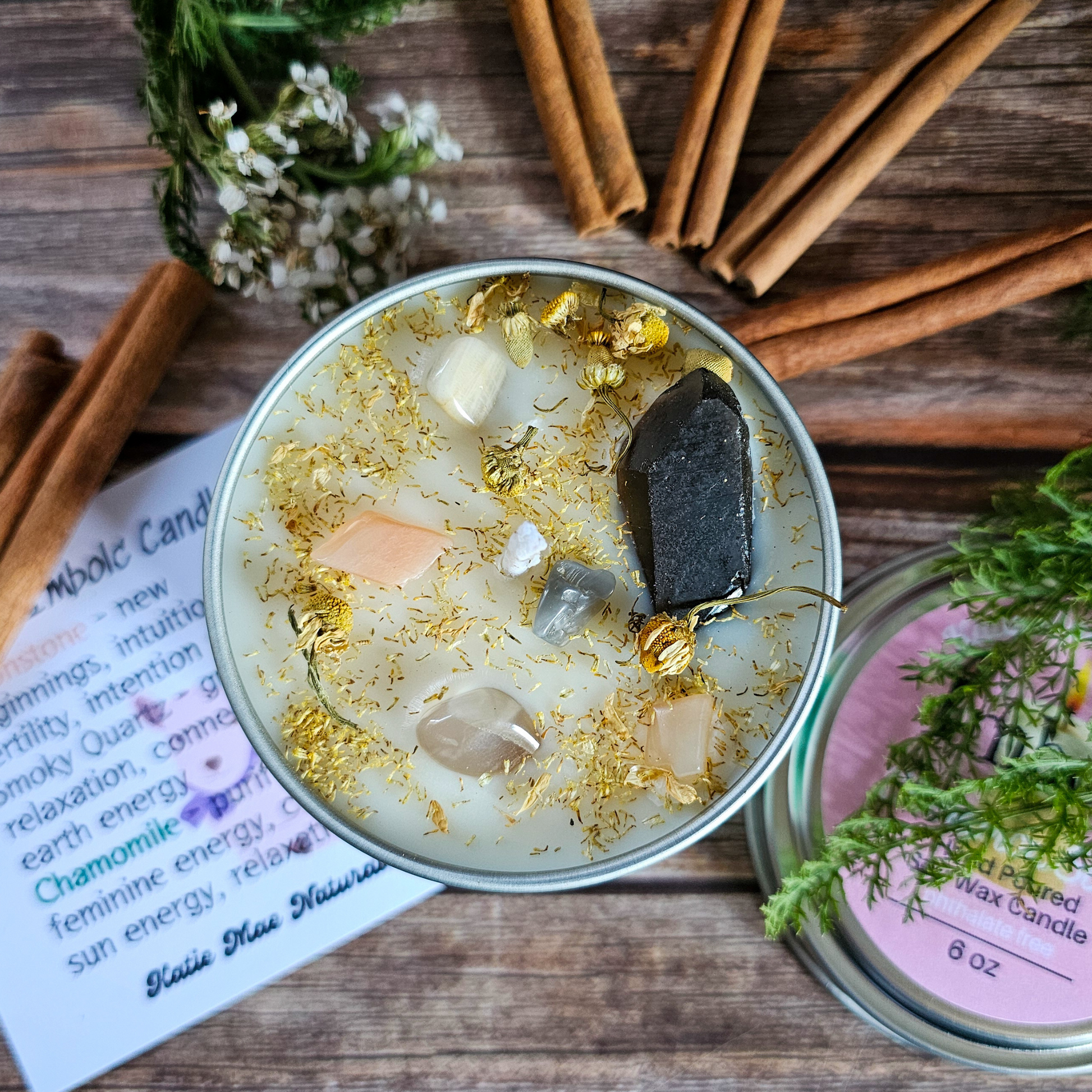 Candle with crystals and herbs on a wooden surface with cinnamon sticks and a label.