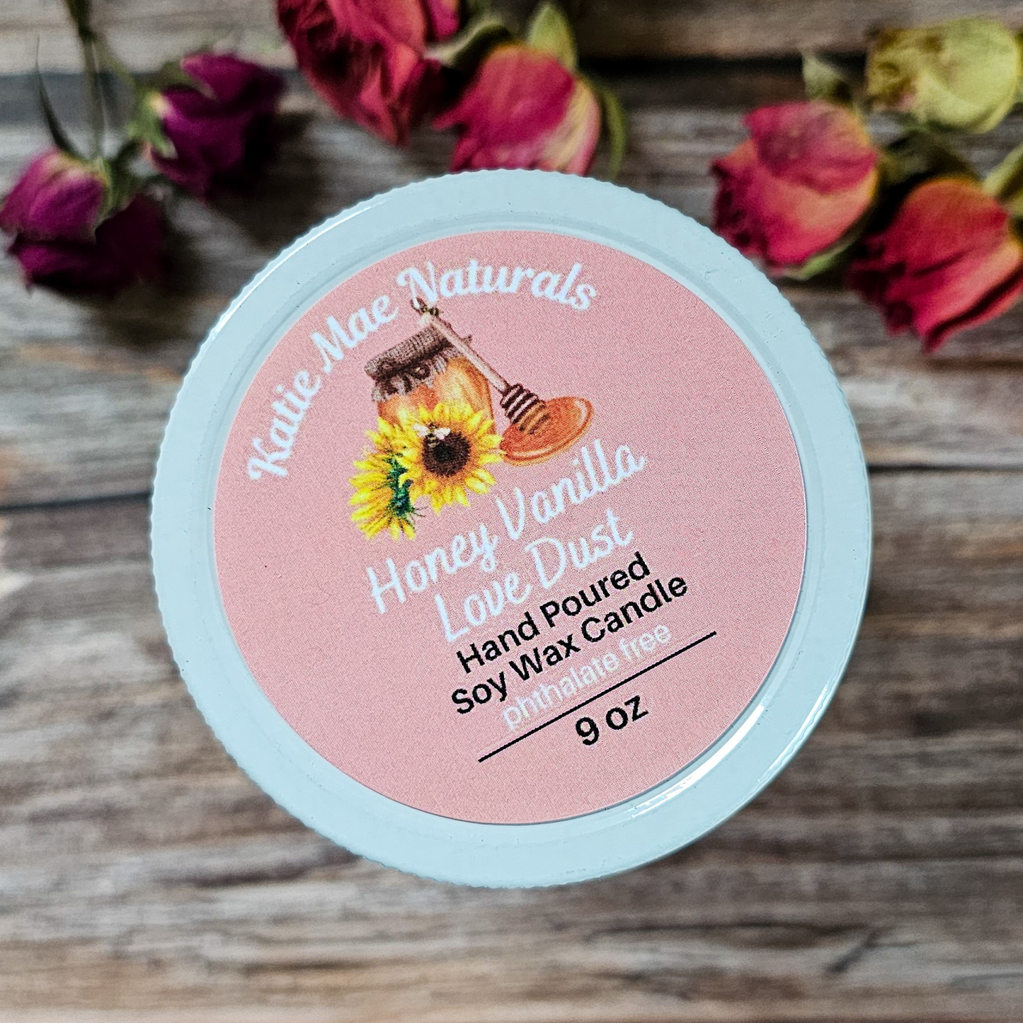 Candle container with pink label featuring 'Katie Mae Naturals' on a wooden surface with flowers.