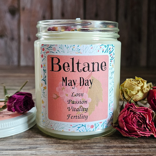 Candle jar with 'Beltane May Day' label surrounded by dried roses on a wooden surface