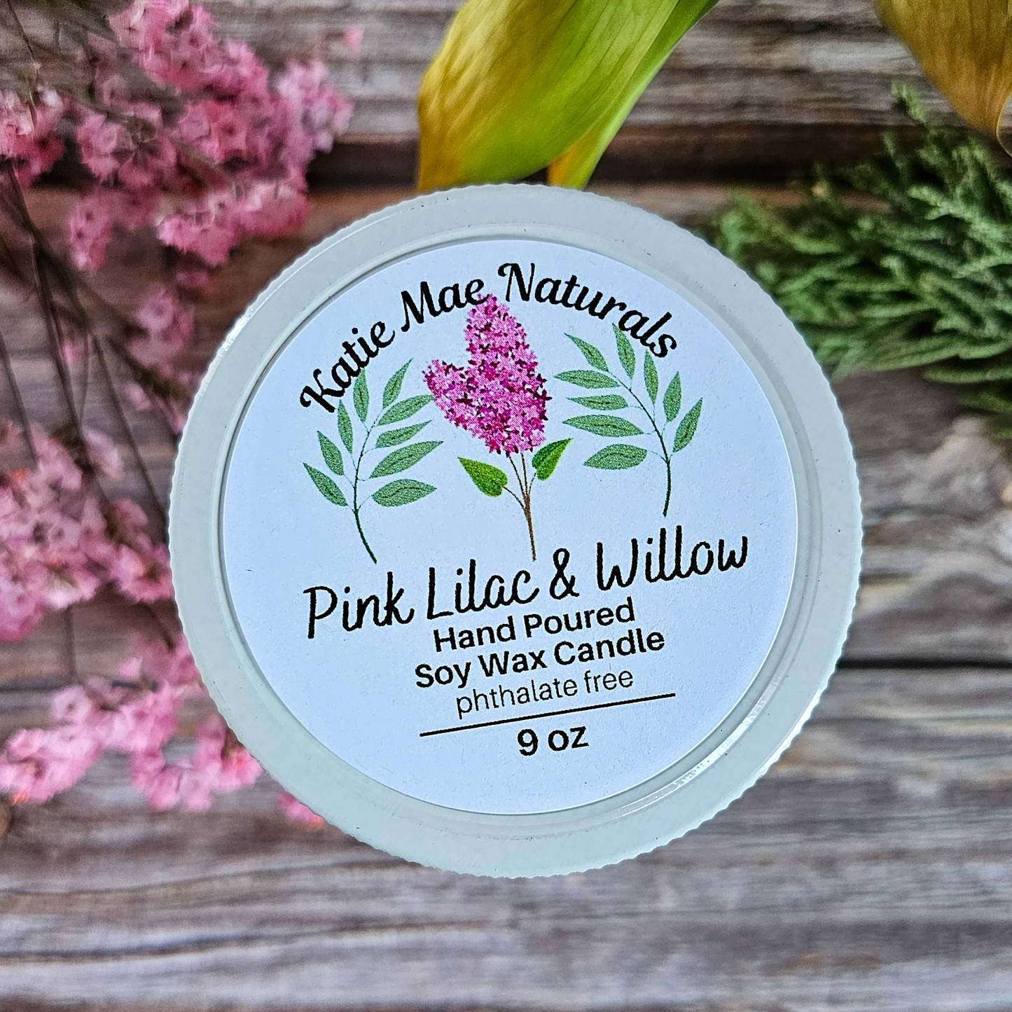 Katie Mae Naturals candle with pink lilac and willow design on a wooden surface with flowers.