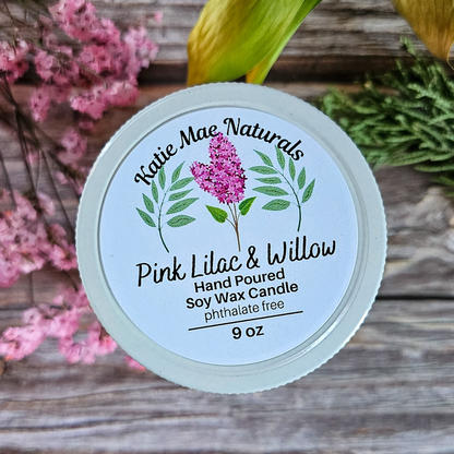 Katie Mae Naturals candle with pink lilac and willow design on a wooden surface with flowers.