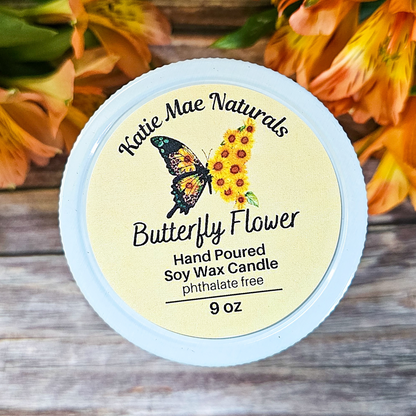 Katie Mae Naturals Butterfly Flower candle with a butterfly and sunflower design on a wooden surface with flowers in the background.