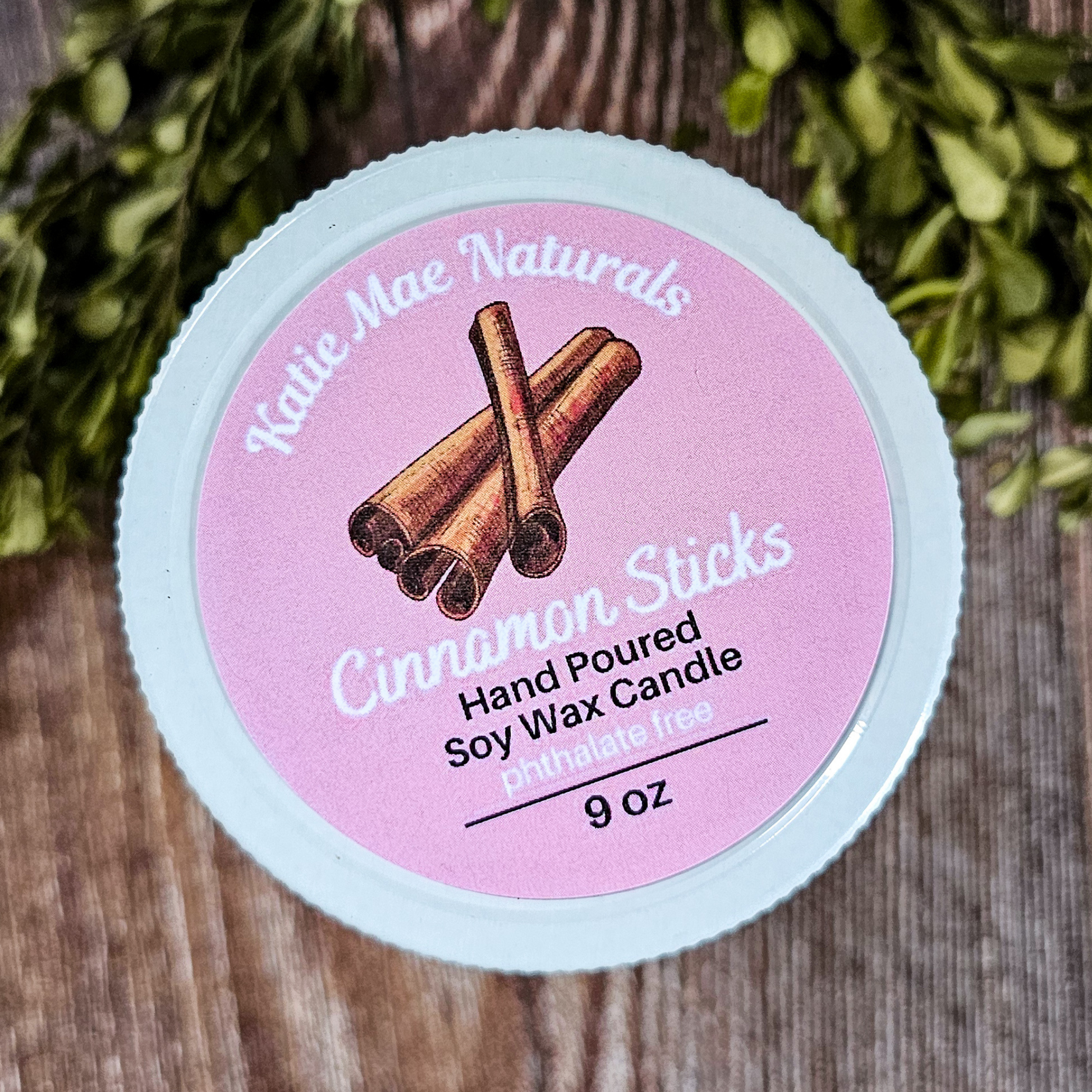 Candle container with 'Katie Mae Naturals' branding and cinnamon stick design on a wooden surface.
