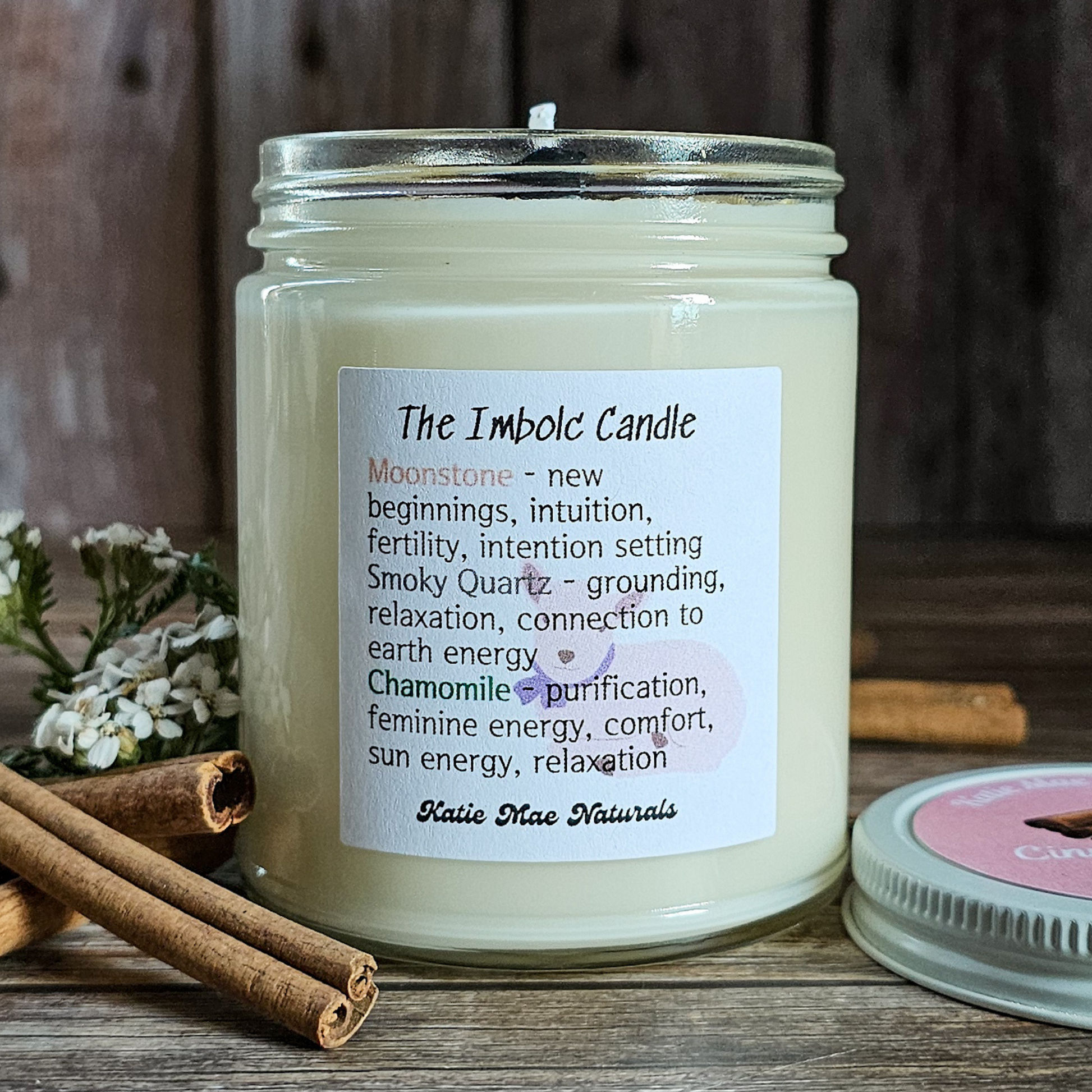 Candle with a label on a wooden surface with cinnamon sticks and flowers.