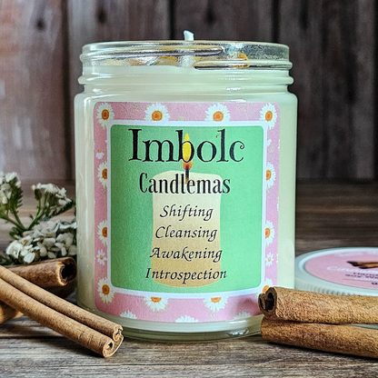 Candle with 'Imbolc Candlemas' label on a wooden surface with cinnamon sticks and flowers.
