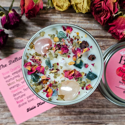 Candle with dried flowers in a tin on a wooden surface with a pink card and flowers in the background.