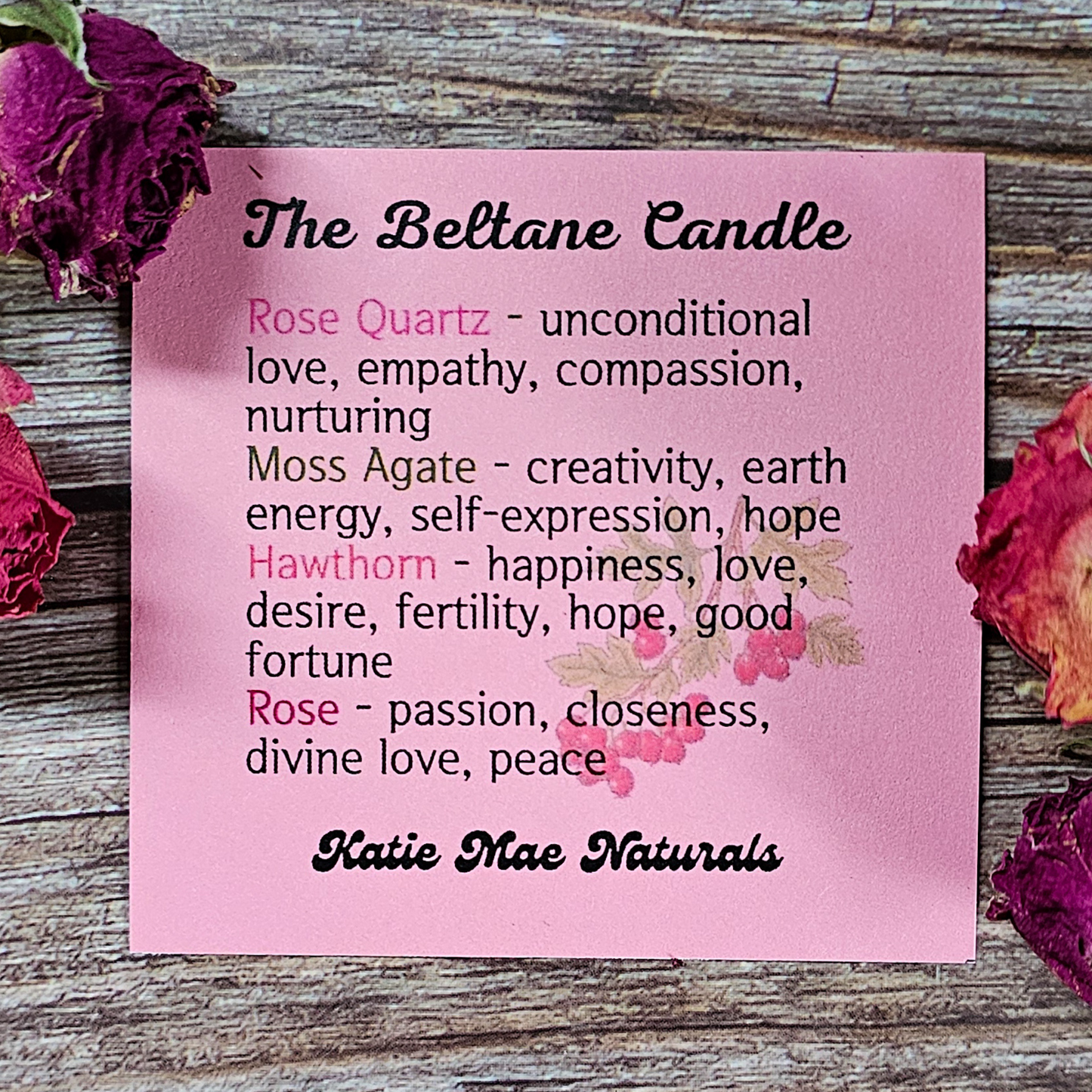 Pink card with text about Beltane candles and their properties, surrounded by dried roses on a wooden surface.