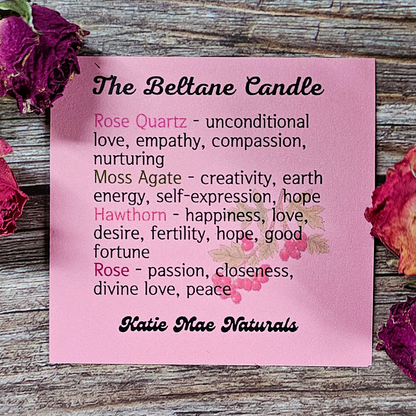 Pink card with text about Beltane candles and their properties, surrounded by dried roses on a wooden surface.