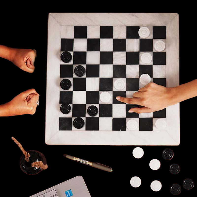 Two people playing checkers on a wooden table with checkers and pieces scattered around.