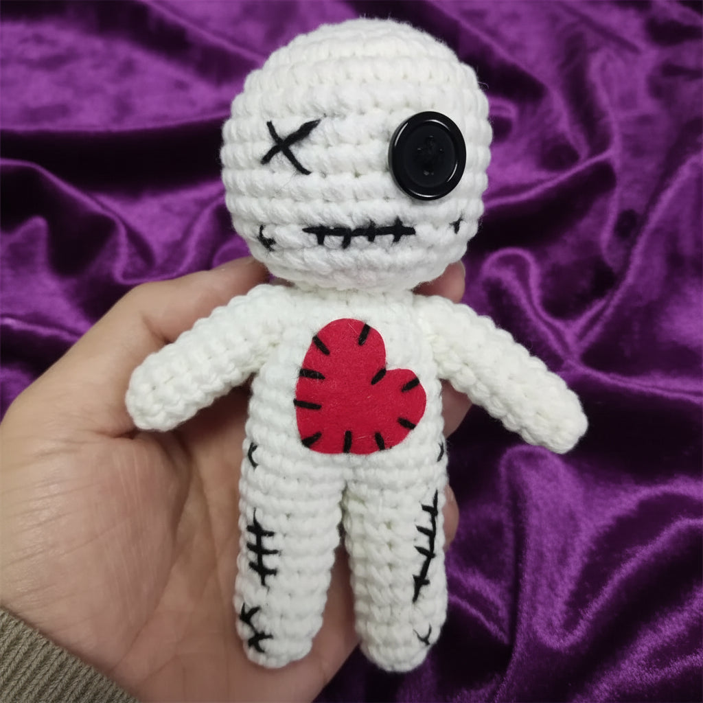 A handmade white crocheted voodoo doll being held in a hand. The doll features a black button for one eye, black stitched details for the other eye and mouth, and a red felt heart on its chest.