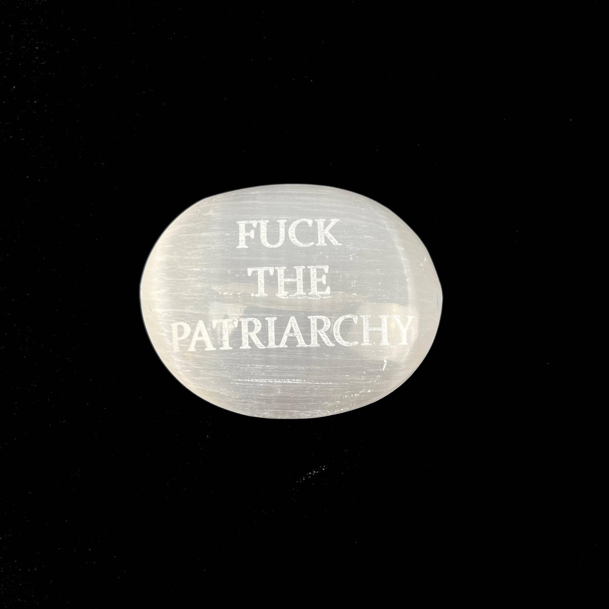 A polished white selenite gemstone with the text 'FUCK THE PATRIARCHY' engraved on it, set against a solid black background.