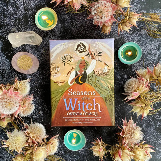 Book titled 'Seasons of the Witch Ostara Oracle' surrounded by candles and flowers on a dark surface